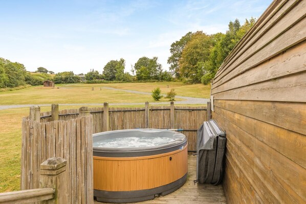 Une terrasse en bois avec un jacuzzi et un espace barbecue couvert, le tout entouré d’une clôture en bois et d’une pelouse.