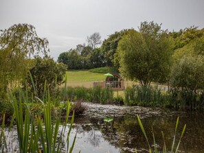 A rustic wooden structure with a green roof, surrounded by lush greenery and a pond.