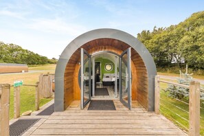 A modern, circular structure with a wooden interior and a glass entrance, leading to a bed and a small seating area.