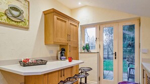 Cocina con armarios de madera, cafetera y una taza roja sobre la encimera. Hay dos taburetes de bar y una mesita.