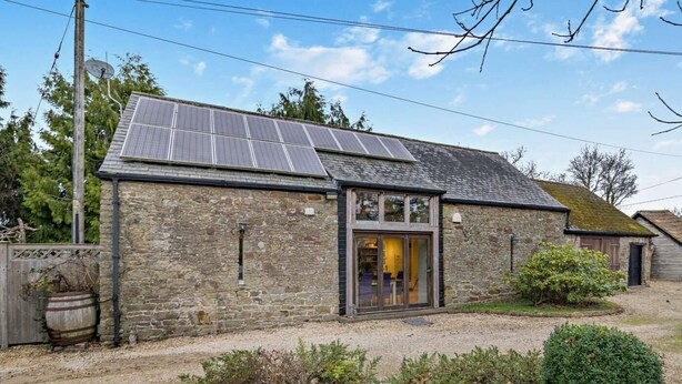 Un edificio de piedra con techo de paneles solares, un barril de madera y un camino de grava.