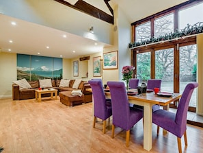 A spacious living area with a wooden dining table, purple chairs, a brown leather sofa, and a large window with a view of trees. - 2 Bed in Coleford (oc-78181) (English Bicknor)