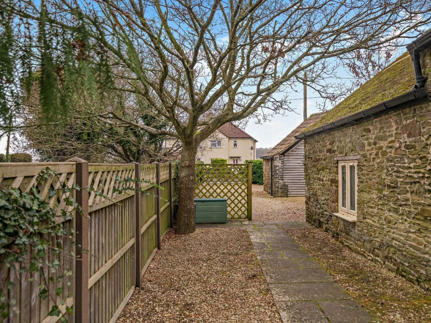 A narrow pathway leading to a stone building with a window, surrounded by a wooden fence and a tree.