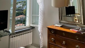 A room with a large window, a wooden dresser with drawers, a mirror, a lamp, and a television on a glass shelf.