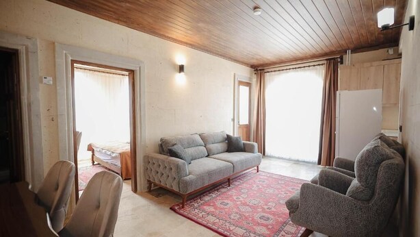 A living room with a sofa, armchairs, a patterned rug, wooden ceiling, and a bed in the background.
