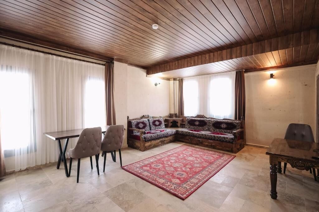 A room with a wooden ceiling, a dining table with chairs, a sofa with patterned cushions, and a red rug.