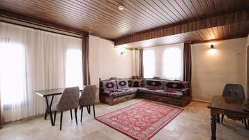 A room with a wooden ceiling, a dining table with chairs, a sofa with patterned cushions, and a red rug.