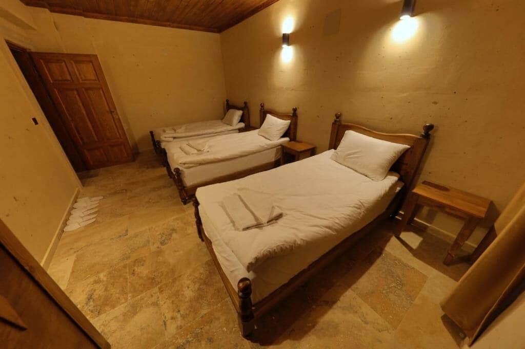 Una habitación con tres camas de madera, cada una con cabecero y mesita de noche de madera. El piso es de baldosas y la puerta es de madera.