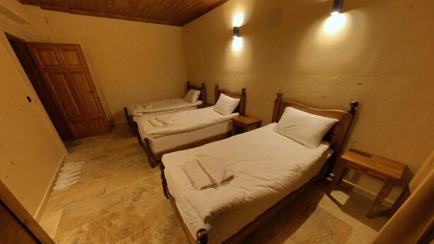 Una habitación con tres camas de madera, cada una con cabecero y mesita de noche de madera. El piso es de baldosas y la puerta es de madera.