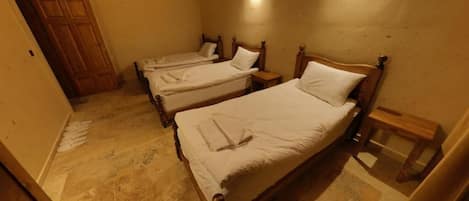 A room with three wooden beds, each with a wooden headboard and a nightstand. The room has a tiled floor and a wooden door.