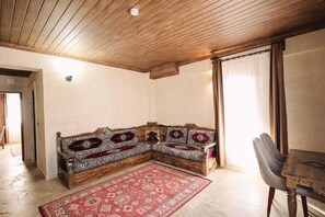 A room with a wooden ceiling, a corner sofa with patterned cushions, a red and white rug, and a dining table with chairs.