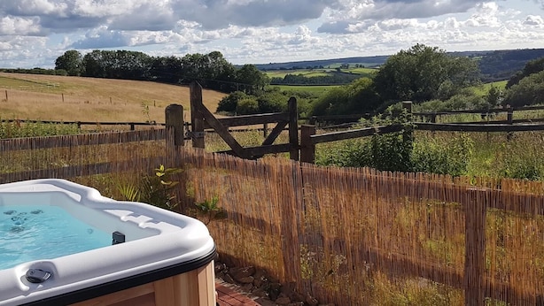 Un bain à remous situé dans un espace extérieur clos, offrant une vue sur des collines verdoyantes et un ciel partiellement nuageux.