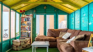Une pièce chaleureuse avec un canapé marron, une petite table, une bibliothèque et une fenêtre donnant sur l’extérieur, où l’on aperçoit un vélo.