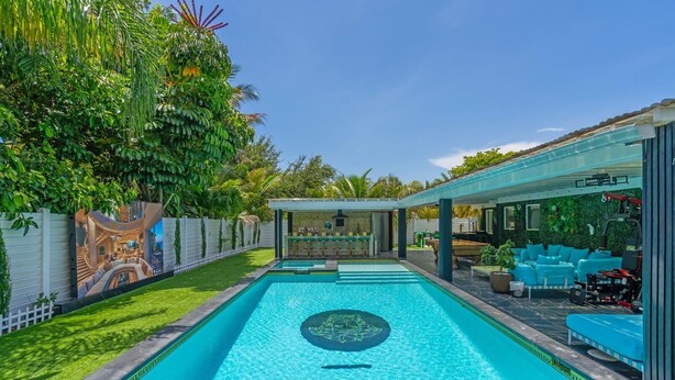 Una piscina rectangular con una isla central, rodeada por un porche cubierto con asientos y un bar.