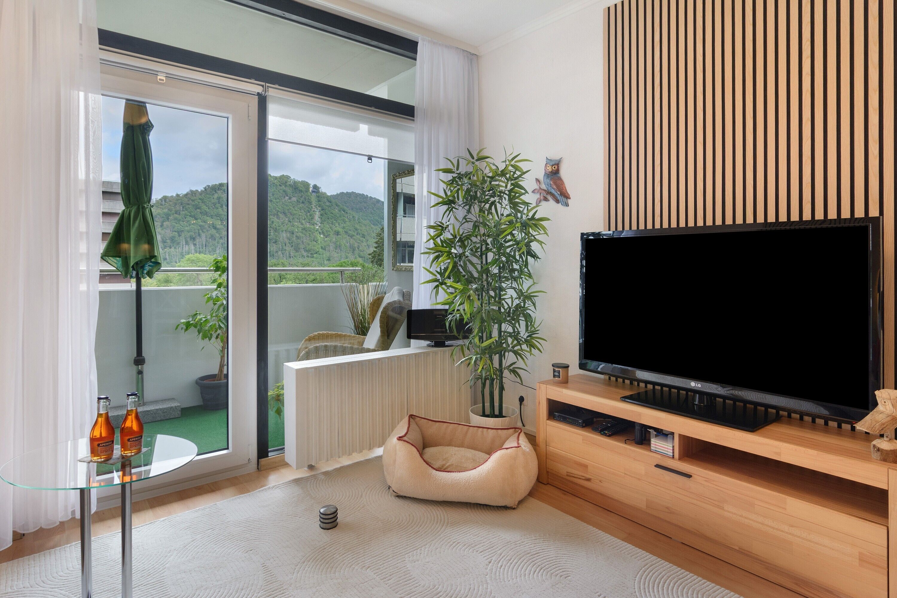 Ein modernes Wohnzimmer mit einem großen Flachbildfernseher, einem bequemen Sitzsack, einer Topfpflanze und Blick auf einen Balkon mit Bergblick.