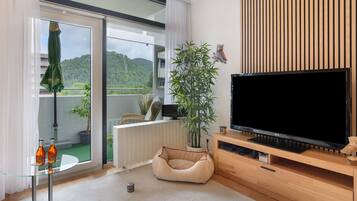 Ein modernes Wohnzimmer mit einem großen Flachbildfernseher, einem bequemen Sitzsack, einer Topfpflanze und Blick auf einen Balkon mit Bergblick.