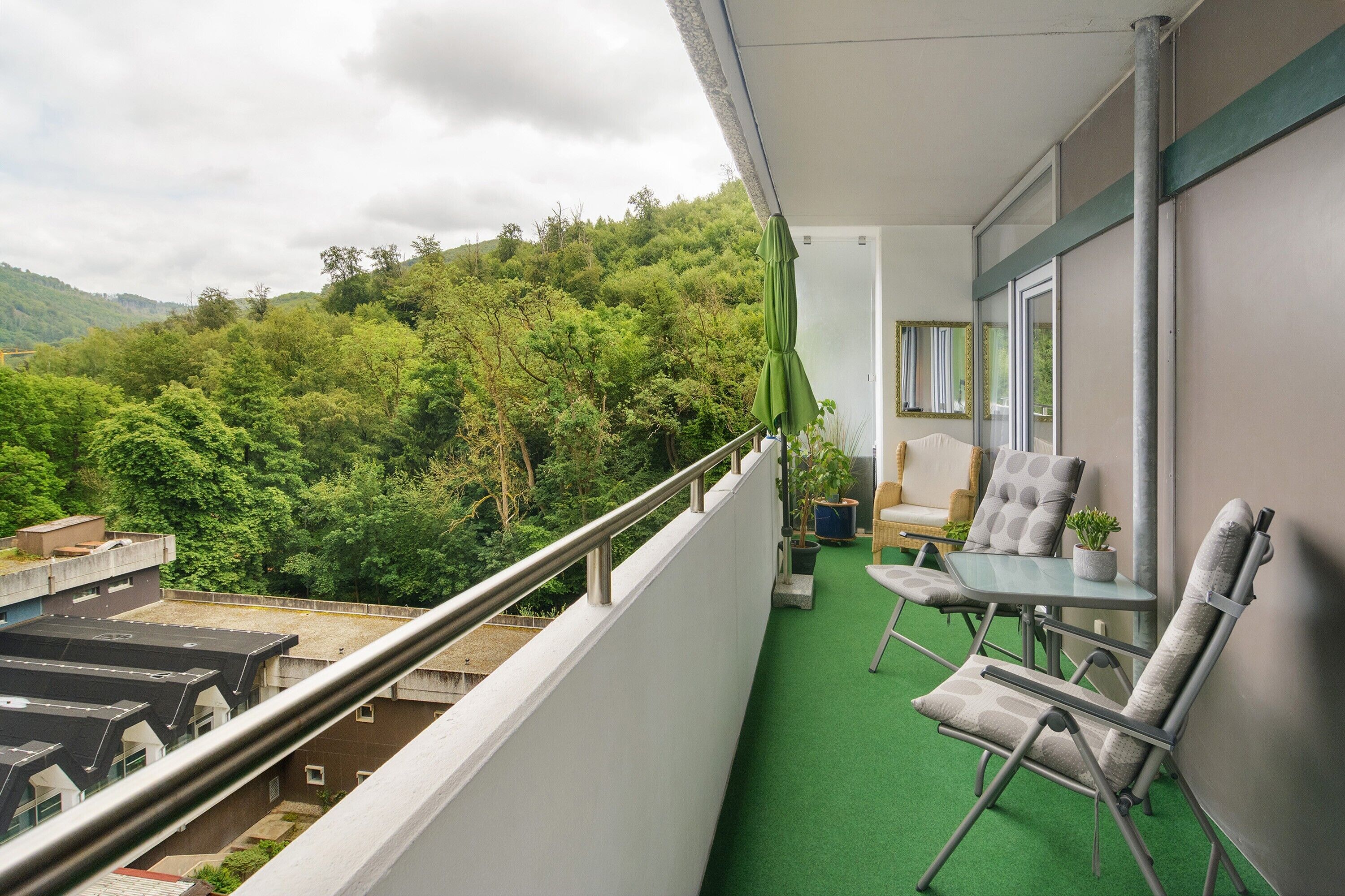 Un balcón con piso de césped artificial verde, una mesa con dos sillas y vistas a una ladera boscosa.