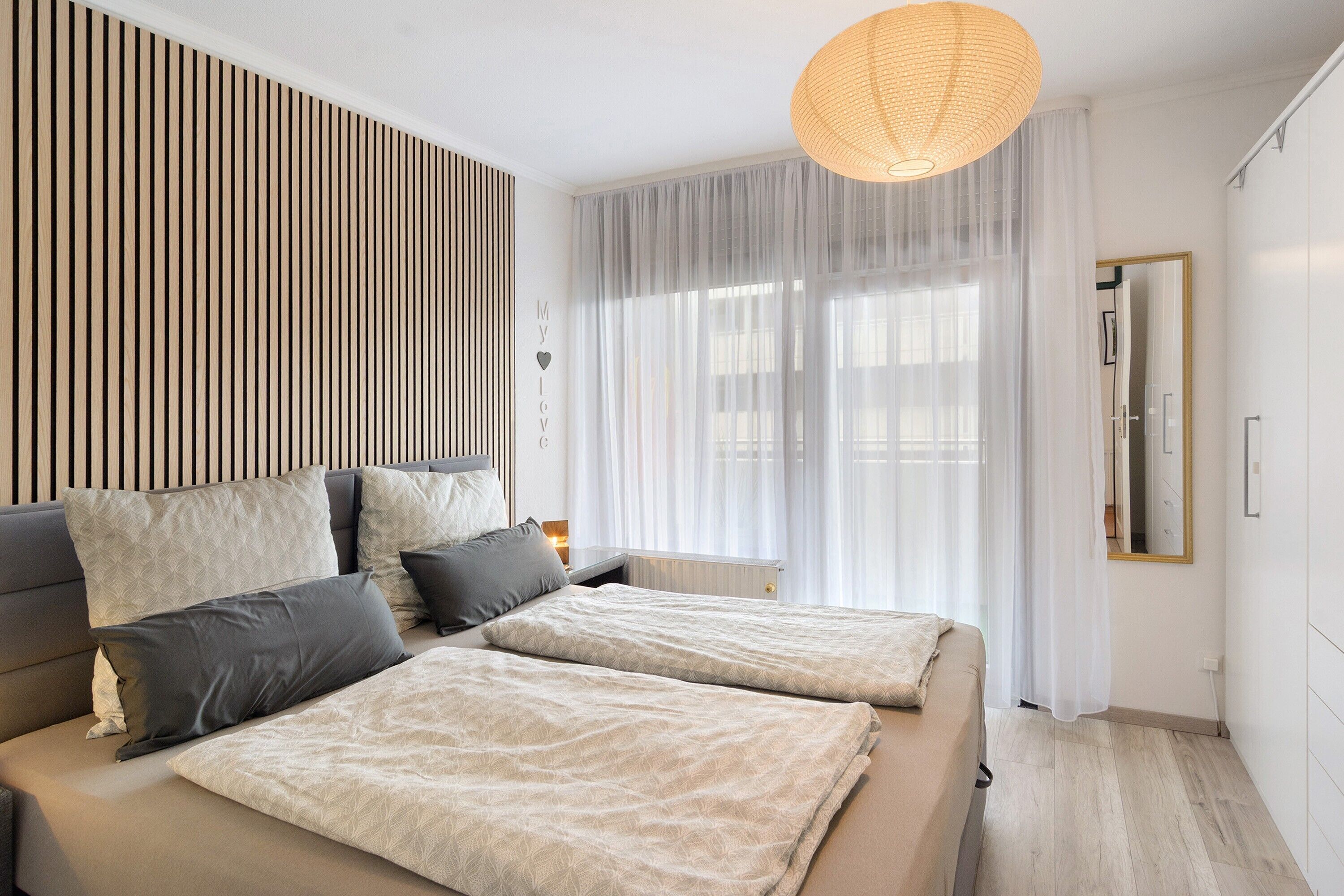 Un dormitorio moderno con cama, mesitas de noche, una lámpara colgante y una ventana con cortinas traslúcidas.