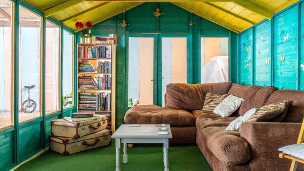 A cozy room with a green ceiling, a brown sofa, a bookshelf, and a window with a bicycle outside. - 3 Bed in Umberleigh (oc-78305) (Kings Nympton)