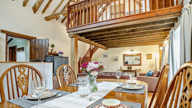 Un comedor con una mesa de madera dispuesta para cuatro, una escalera que sube a un nivel superior y una sala de estar acogedora al fondo.