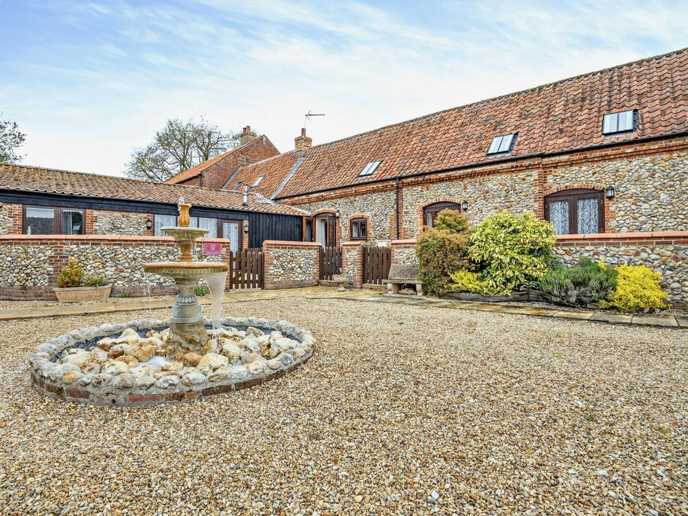 A courtyard with a stone fountain, a gravel ground, and a stone building with a tiled roof.