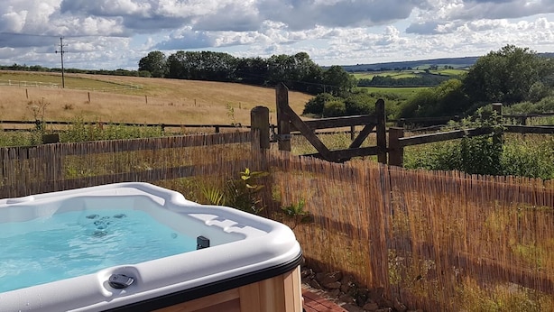 A hot tub with a wooden deck, surrounded by a wooden fence, overlooking a rural landscape with fields and a cloudy sky. - 2 Bed in Umberleigh (oc-78303) (Kings Nympton)