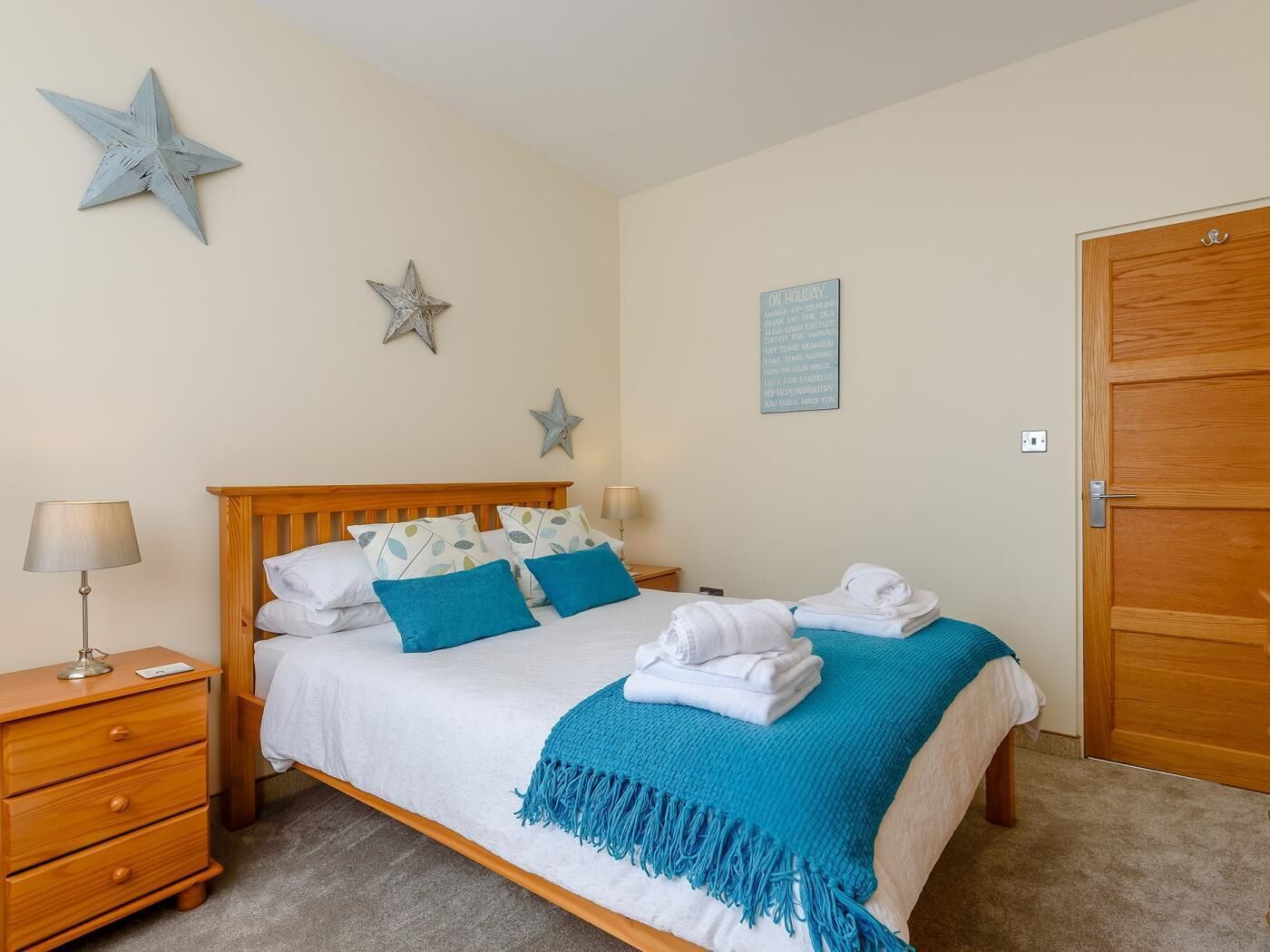 A neatly made bed with a blue blanket and pillows, a wooden nightstand with a lamp, and a wooden door.