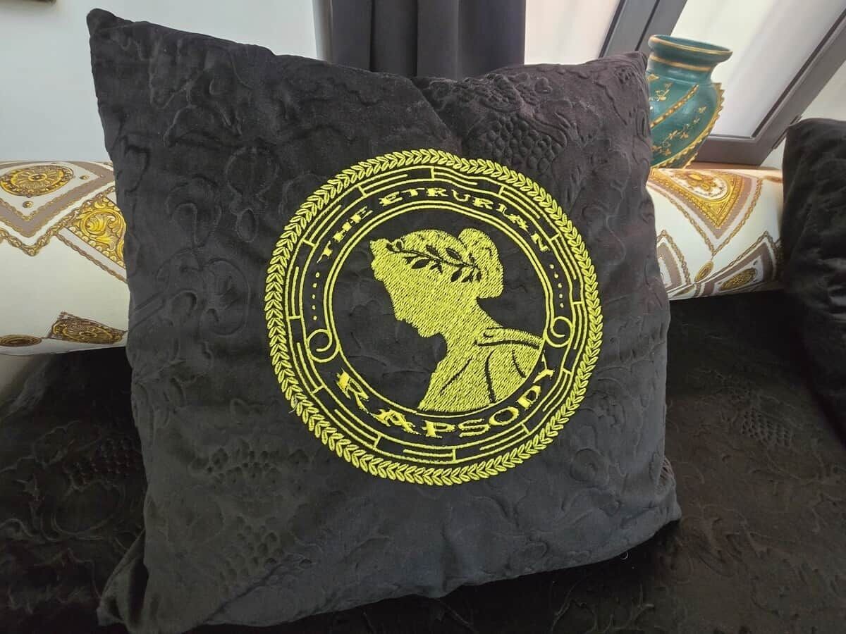 A black pillow with a gold embroidered emblem featuring a profile of a woman and the text 'THE ETRURIAN RAPSODY.'