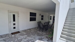 En overdækket terrasse med stengulv, en hvid dør og et bord med stole.