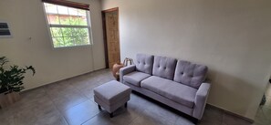 A living room with a grey sofa, a matching ottoman, a potted plant, a window with blinds, and a wooden door. - Come for the beaches? Villa Marcos, unique 2-bedrooms apartment Curacao with AC (Willemstad)
