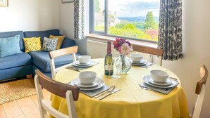 A dining area with a table set for four, a sofa in the background, and a window with a view of greenery. - 2 Bed in Cinderford (oc-65670) (Littledean)