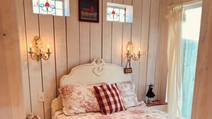 Une chambre à coucher avec une couvre-lit à motifs floraux, une tête de lit en forme de cœur, deux appliques murales, une petite table de chevet ornée d’une figurine et une fenêtre avec un rideau.