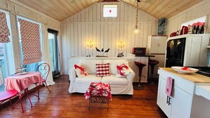 Une pièce chaleureuse avec un plafond en bois, un canapé blanc agrémenté de coussins rouges et blancs, une petite table recouverte d’une nappe à motifs floraux, et un coin cuisine équipé d’un réfrigérateur et d’un four à micro-ondes.