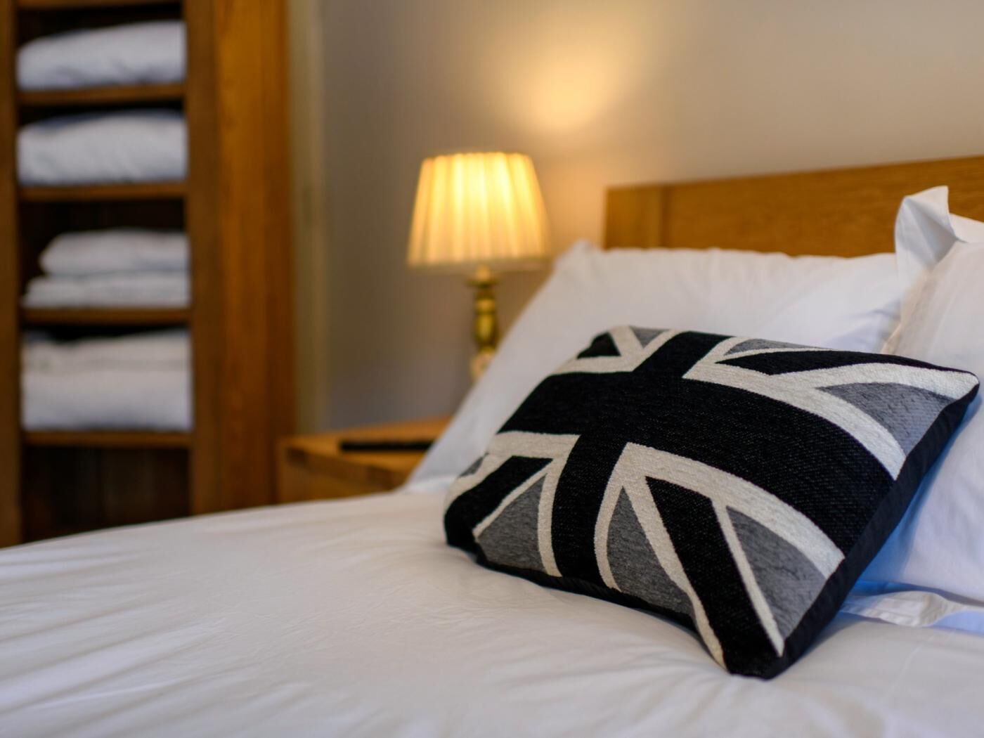 A neatly made bed with white linens, a pillow with a Union Jack design, a wooden headboard, and a bedside lamp.