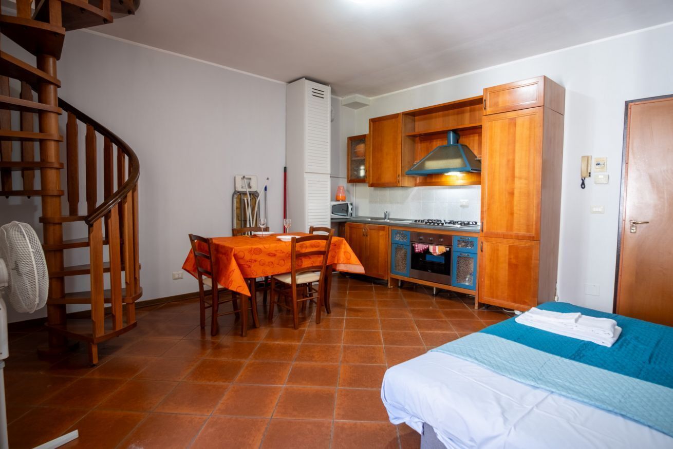 A compact kitchen with wooden cabinets, a dining table with orange tablecloth, a bed with blue bedding, and a spiral staircase.