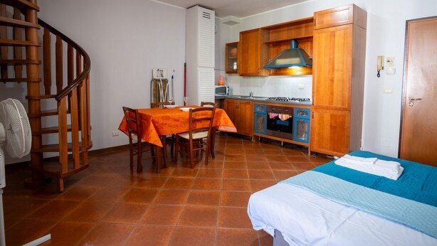 Eine kleine Küche mit Holzschränken, ein Esstisch mit orangefarbener Tischdecke, ein Bett mit blauer Bettwäsche und eine Wendeltreppe.