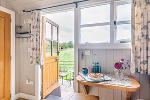 A room with a wooden door leading to an outdoor area, a table with pastries and a vase of flowers, and a view of a grassy landscape.