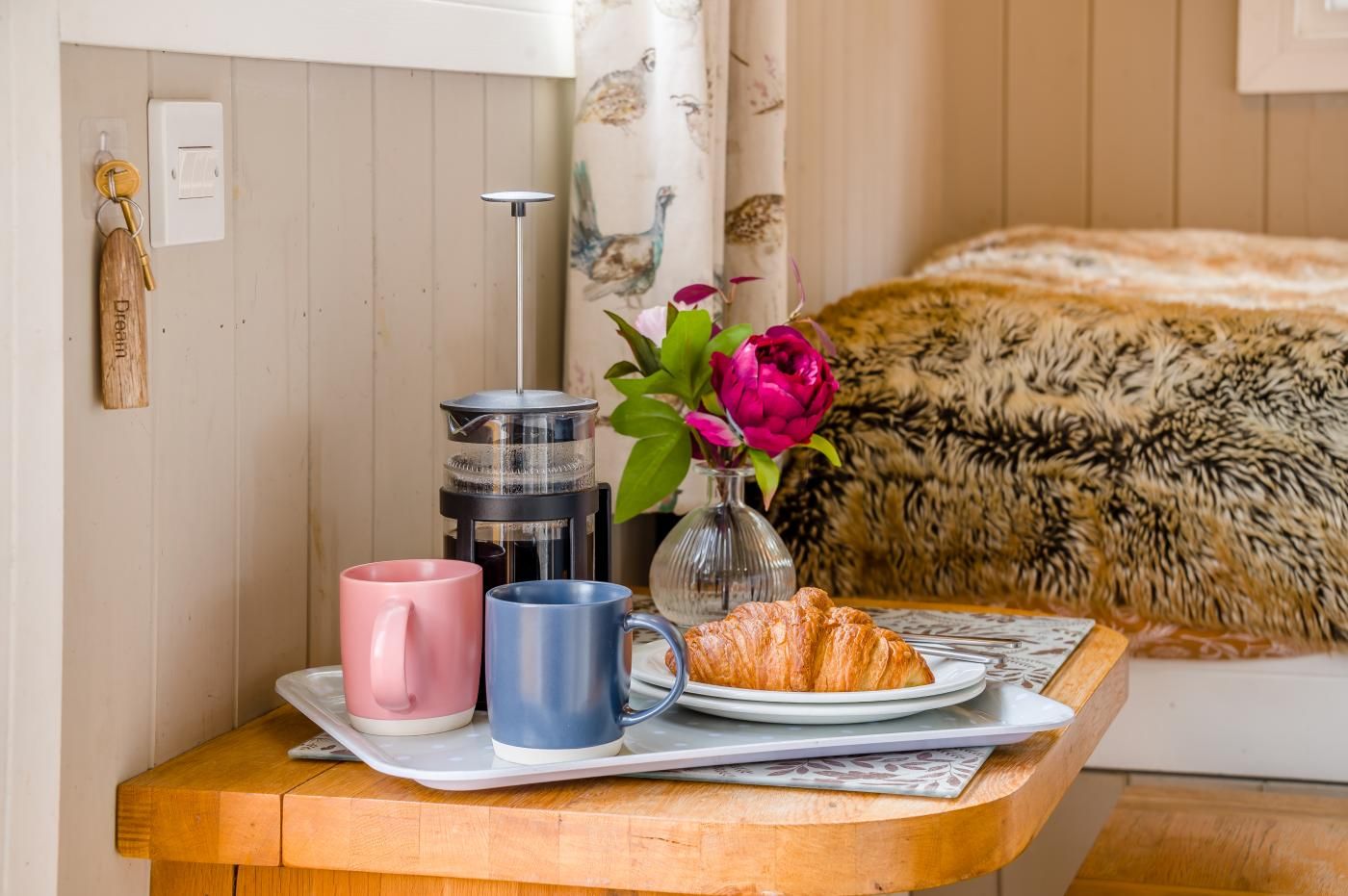 A breakfast setup with a coffee maker, two mugs, a croissant, and a vase with flowers on a wooden table.