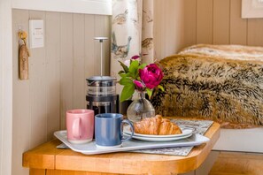 A breakfast setup with a coffee maker, two mugs, a croissant, and a vase with flowers on a wooden table.