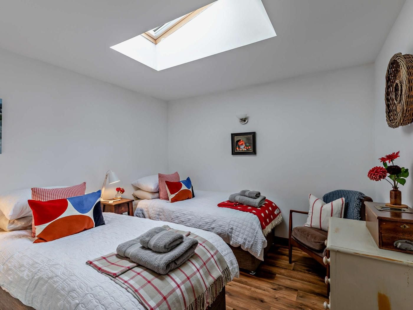 A room with two beds, a skylight, a small table, a chair, and a wall-mounted lamp.