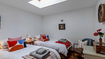 A room with two beds, a skylight, a small table, a chair, and a wall-mounted lamp.