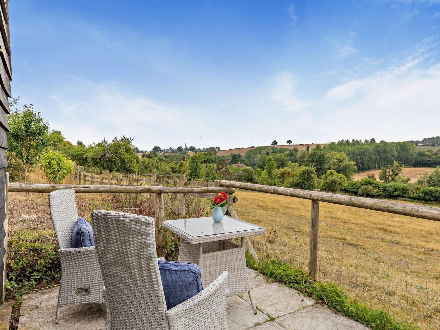 Une terrasse dotée de meubles en osier et d’une table, offrant une vue sur un paysage rural avec des champs et des arbres.