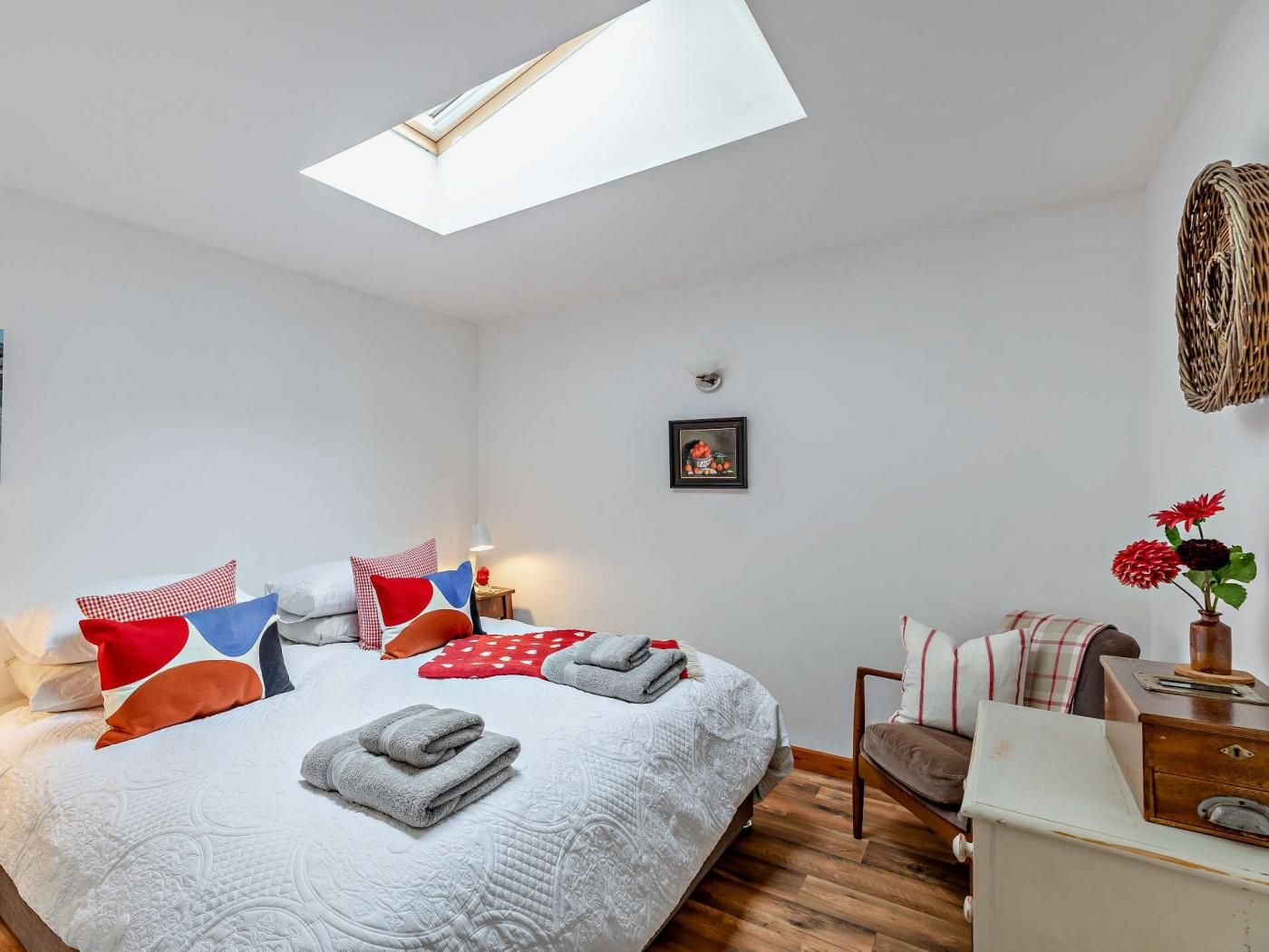 A bedroom with a skylight, a bed with multiple pillows, a bedside table with a lamp, and a dresser with a vase of flowers.