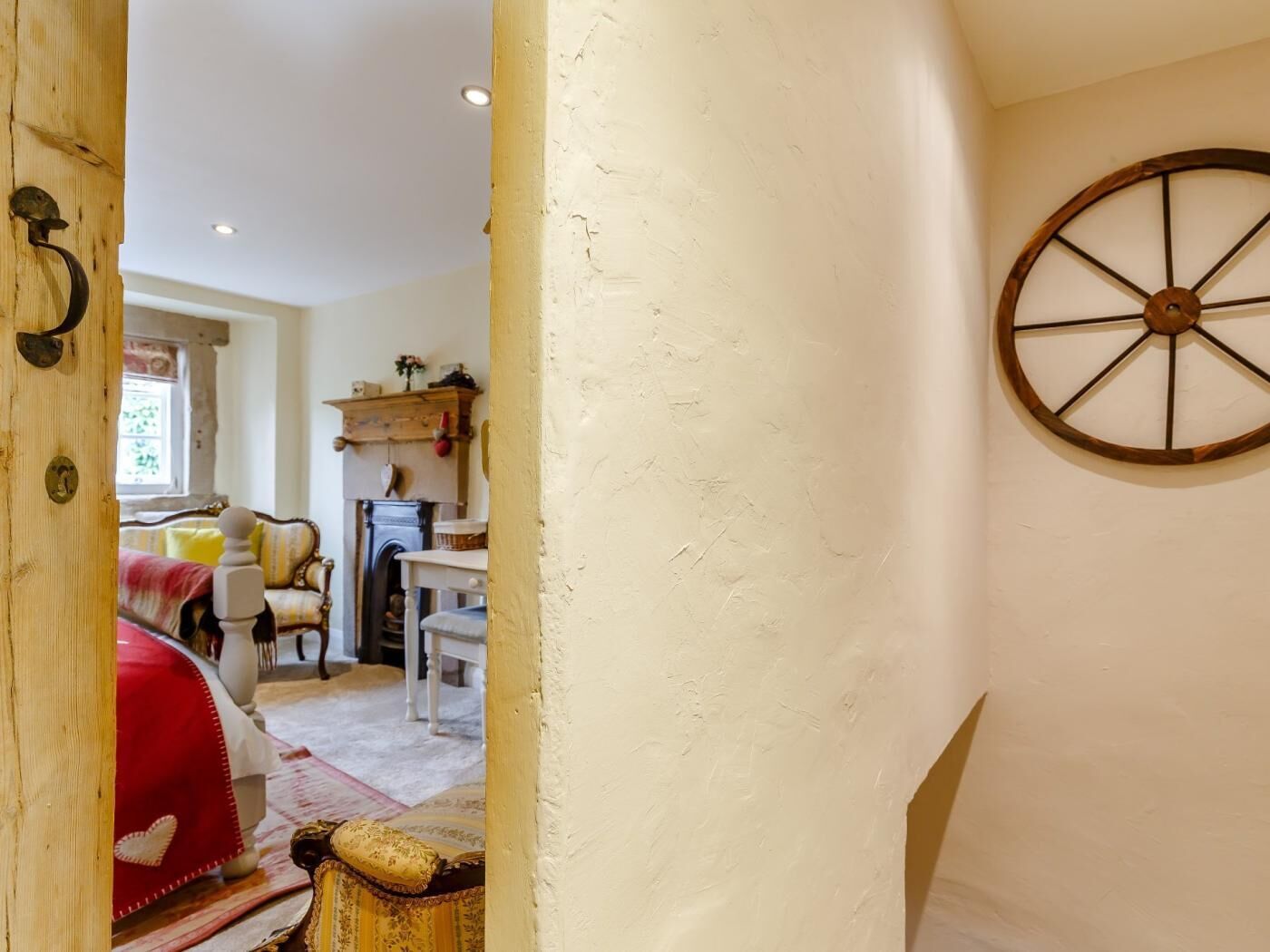 Una habitación con puerta de madera, pared blanca decorada con un detalle de rueda de madera, cama con cubrecama rojo y ventana con cortinas.