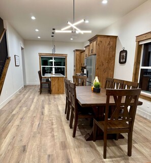 A modern kitchen with wooden cabinets, a stainless steel refrigerator, and a dining area with wooden chairs and a table. - Red Dog Retreat | Hot Tub + A/C, Island Park (Island Park)