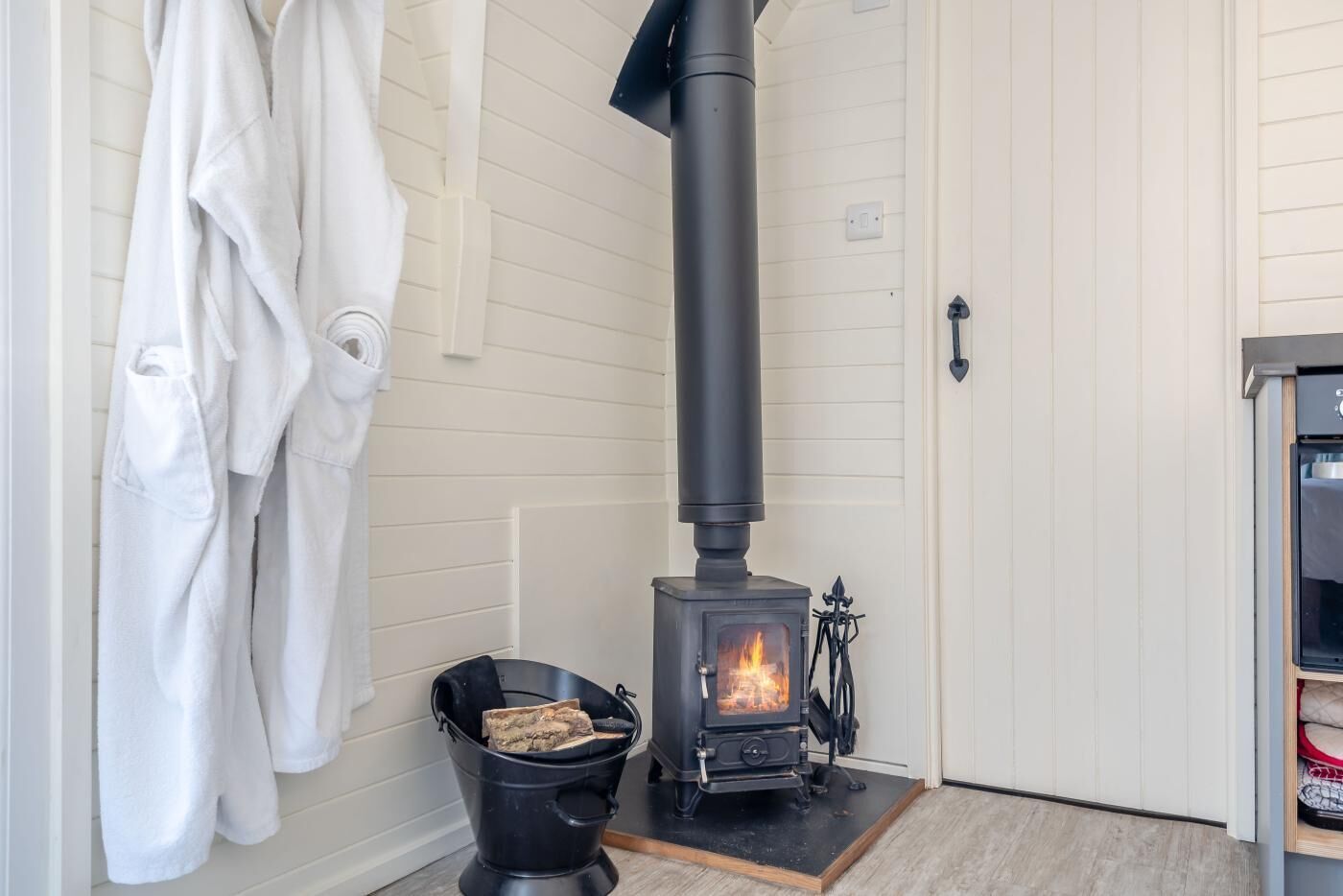 Une pièce avec un poêle à bois, un seau de bois de chauffage et des serviettes blanches accrochées au mur.