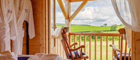 A wooden cabin room with a bed, a chair, and a balcony with a view of the countryside.