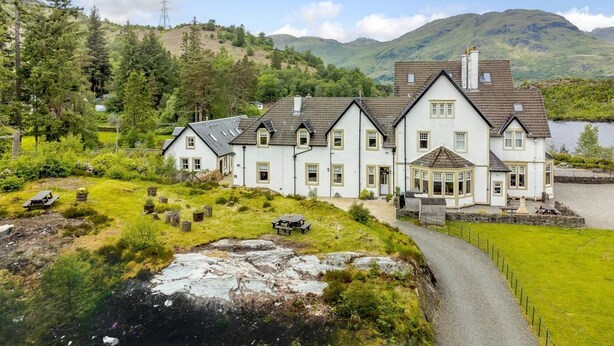 Un grand bâtiment blanc, doté de nombreuses fenêtres et d’une cheminée, entouré de verdure et bordé par un lac.