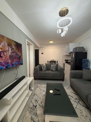 Ein Wohnzimmer mit Flachbildfernseher, einer Couch, einem Couchtisch und einer Küche im Hintergrund.