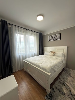 Ein Schlafzimmer mit einem Bett, einem fenster mit Vorhängen, einer Deckenlampe, einem Wandbild und einem Teppich mit Marmormuster.